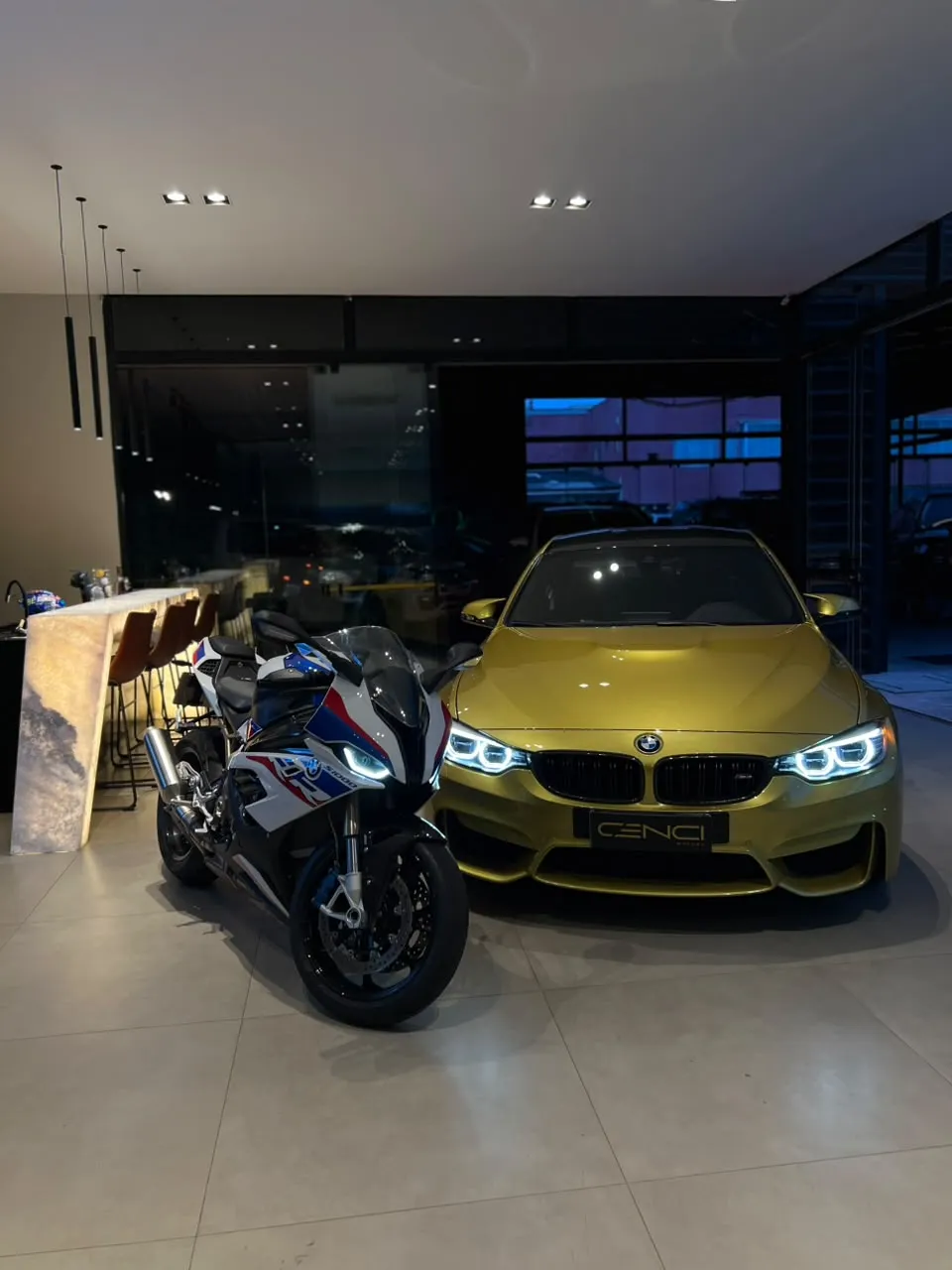 BMW M4 amarela de faróis acesos ao lado de uma moto BMW S 1000 RR no salão da Cenci Motors
