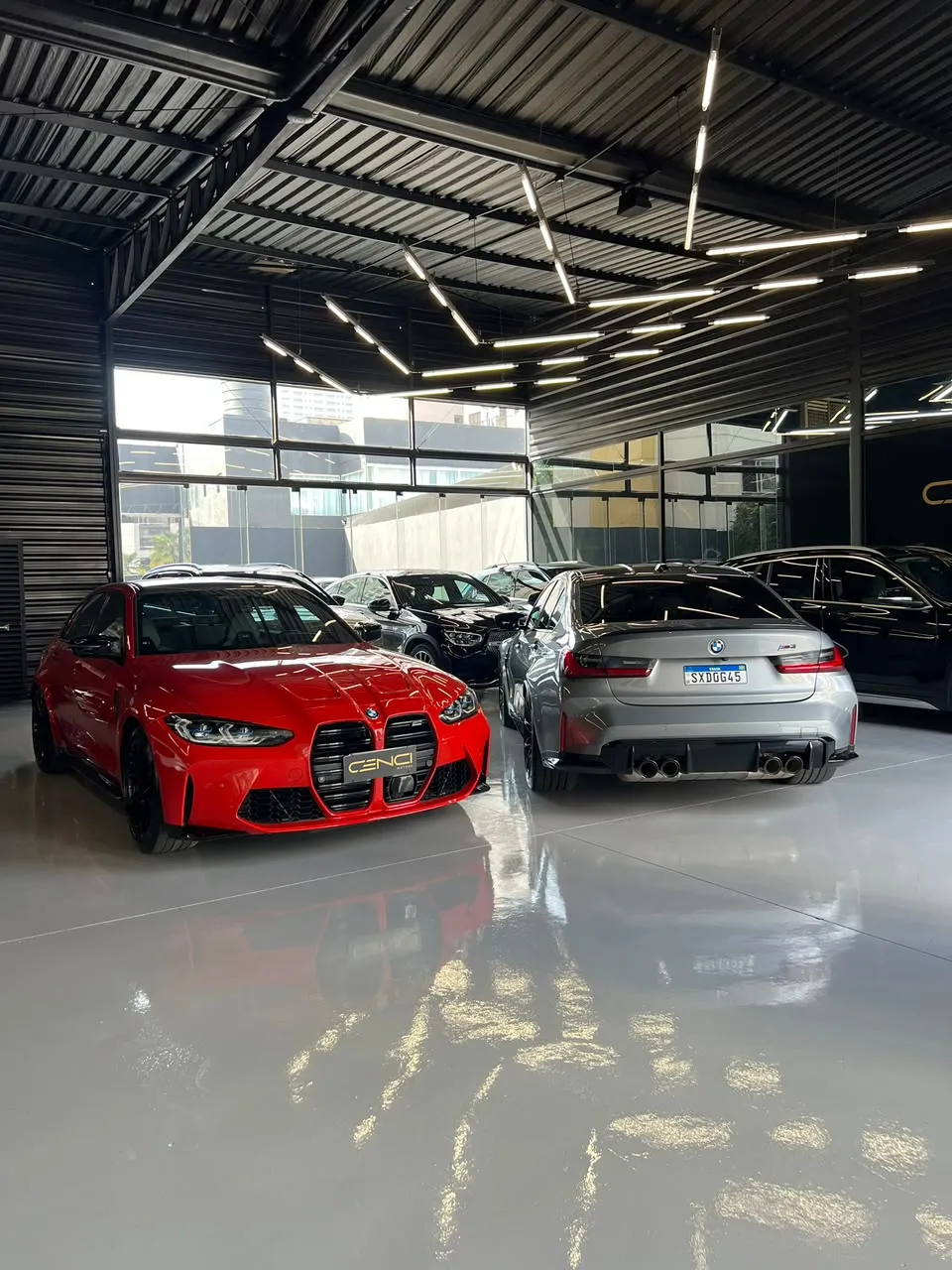 BMW M3 vermelha e BMW M3 cinza sobre o piso claro que reflete as luzes do teto